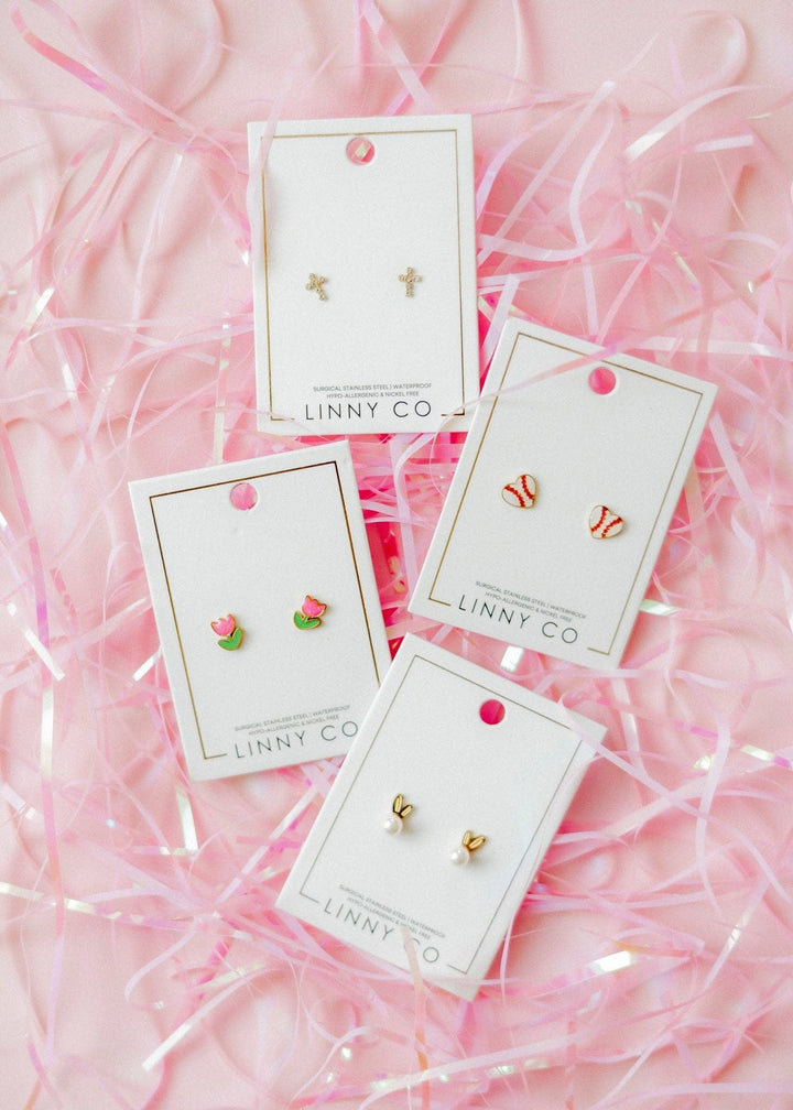 Four Linny Co stud earring cards flat lay on pink ribbon confetti background — gold cross studs, pink tulip enamel studs, red and white baseball enamel studs, and gold heart studs.