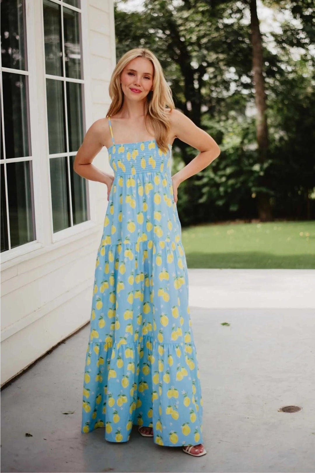 Smiling woman with hand on hip wearing a light blue lemon print smocked tiered maxi dress with spaghetti straps, standing on a patio with greenery in the background.