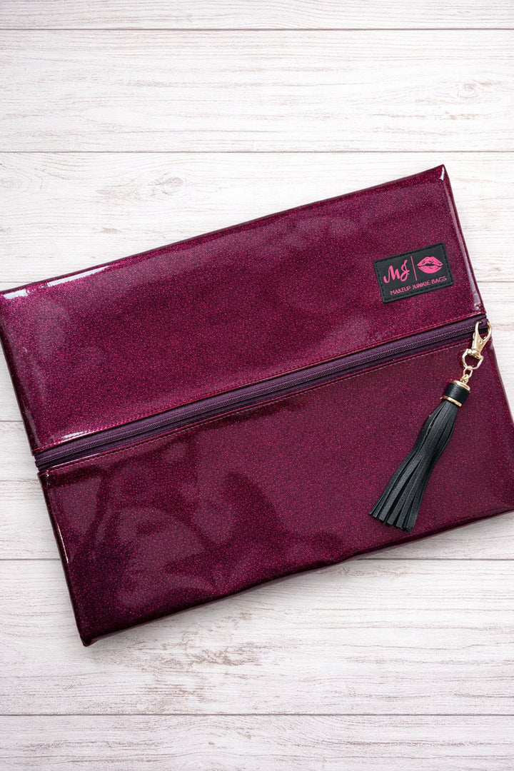 Makeup Junkie Bags magenta hot pink glitter foldover zip makeup bag closed with black tassel zipper pull and branded patch, front view on white wood background