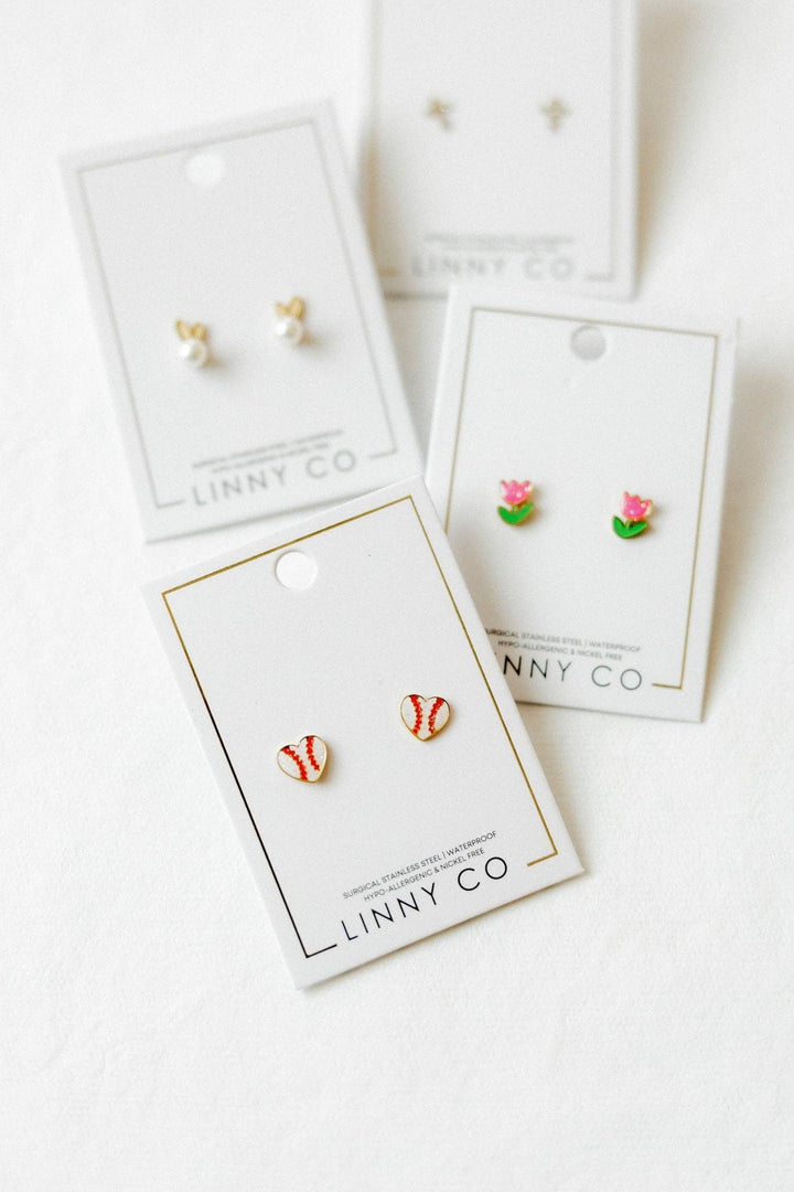 Linny Co stud earring cards flat lay on white background — red and white baseball enamel studs in foreground, with gold bunny ear pearl studs, pink tulip enamel studs, and gold cross stud cards partially visible behind.
