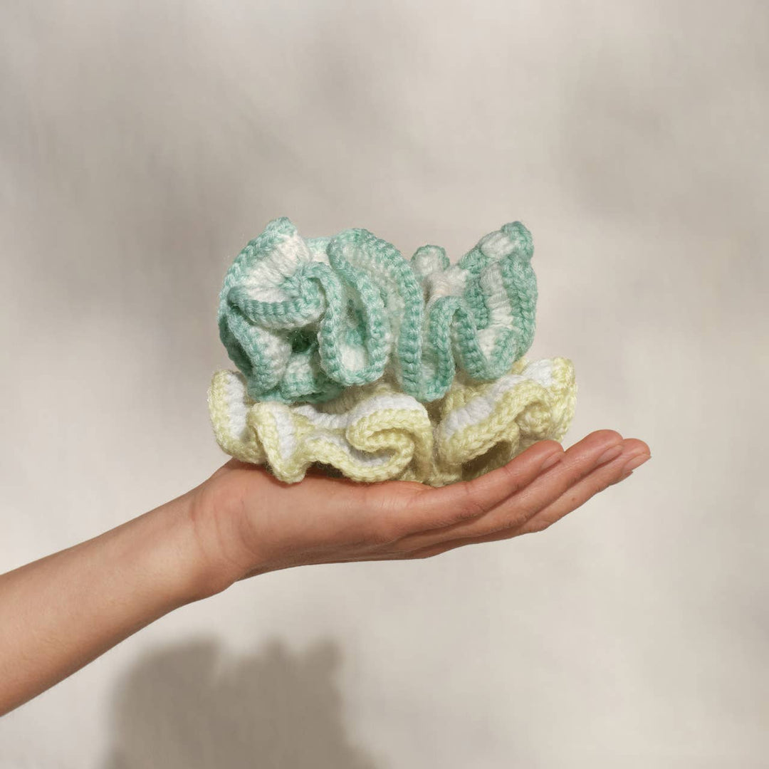 Hand holding two oversized crochet scrunchies — one mint green and white stripe and one butter yellow — stacked on an open palm against a neutral background.