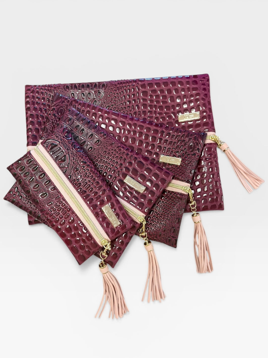Four-piece burgundy crocodile embossed faux leather bag set — large fold-over clutch, medium zip pouch, small zip pouch, and mini zip pouch — all with gold zippers and blush pink tassel pulls, on a white background.