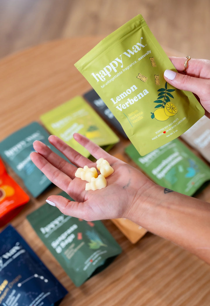 Hand holding Happy Wax Lemon Verbena pouch and yellow bear-shaped wax melts over a wood surface with assorted Happy Wax pouches.