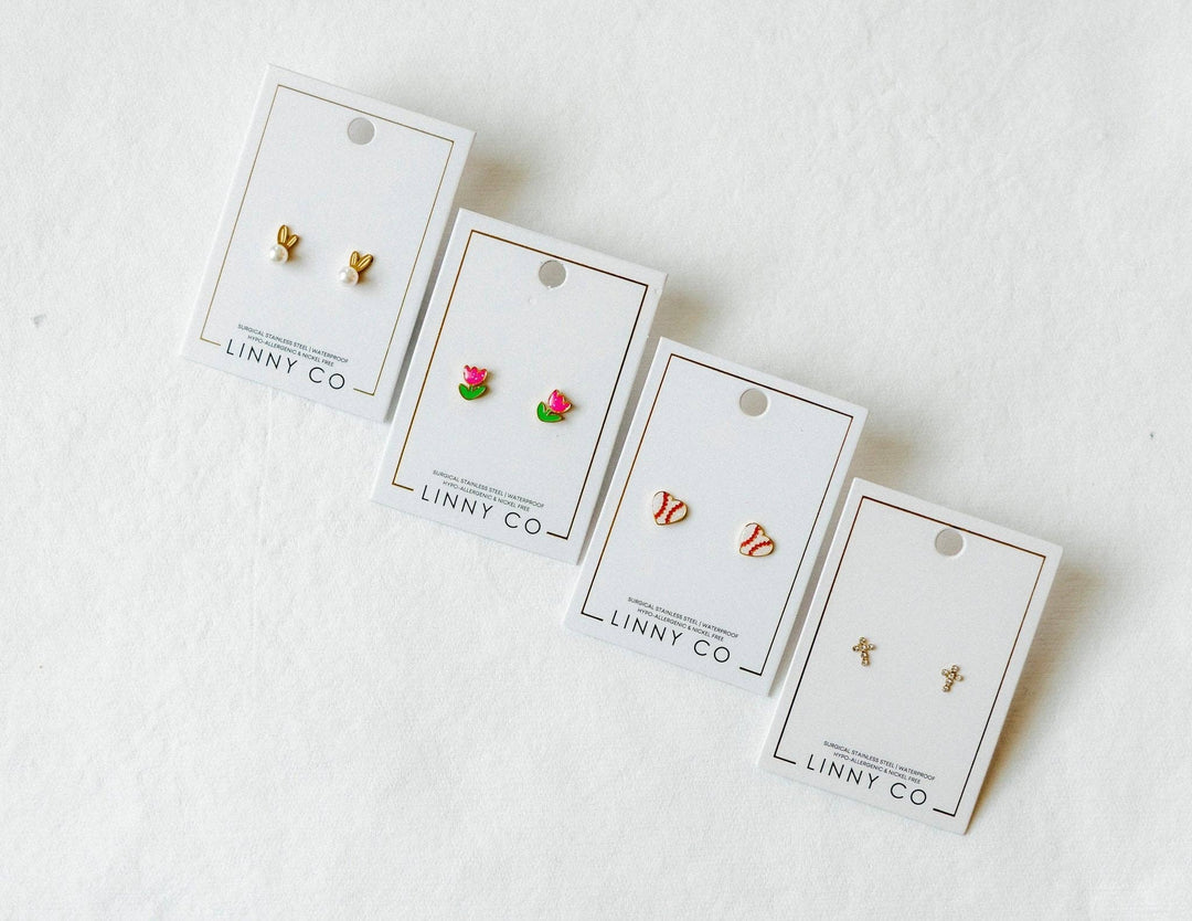 Four Linny Co stud earring cards in a row on white background — gold bunny ear pearl studs, pink tulip enamel studs, red and white baseball enamel studs, and gold cross studs.