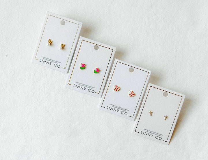 Four Linny Co stud earring cards in a row on white background — gold bunny ear pearl studs, pink tulip enamel studs, red and white baseball enamel studs, and gold cross studs.