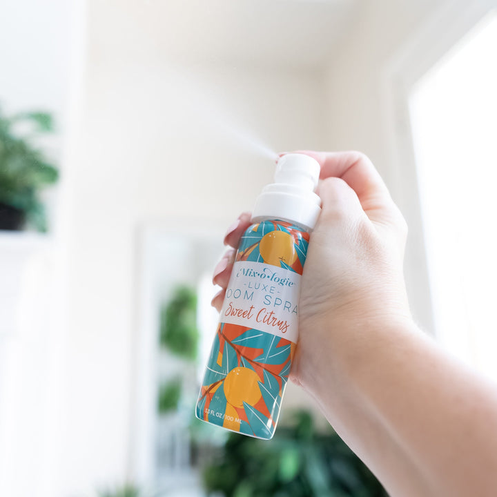 Mixologie Luxe Room Spray in Sweet Citrus, 3.3 fl oz, clear pump bottle with teal and orange citrus illustrated label, held mid-spray in a bright room