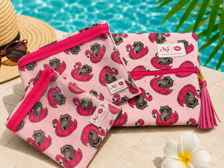 Makeup Junkie Bags set of three pink cosmetic bags with raccoon on flamingo pool float print and hot pink zipper, shown poolside with straw hat and sunglasses