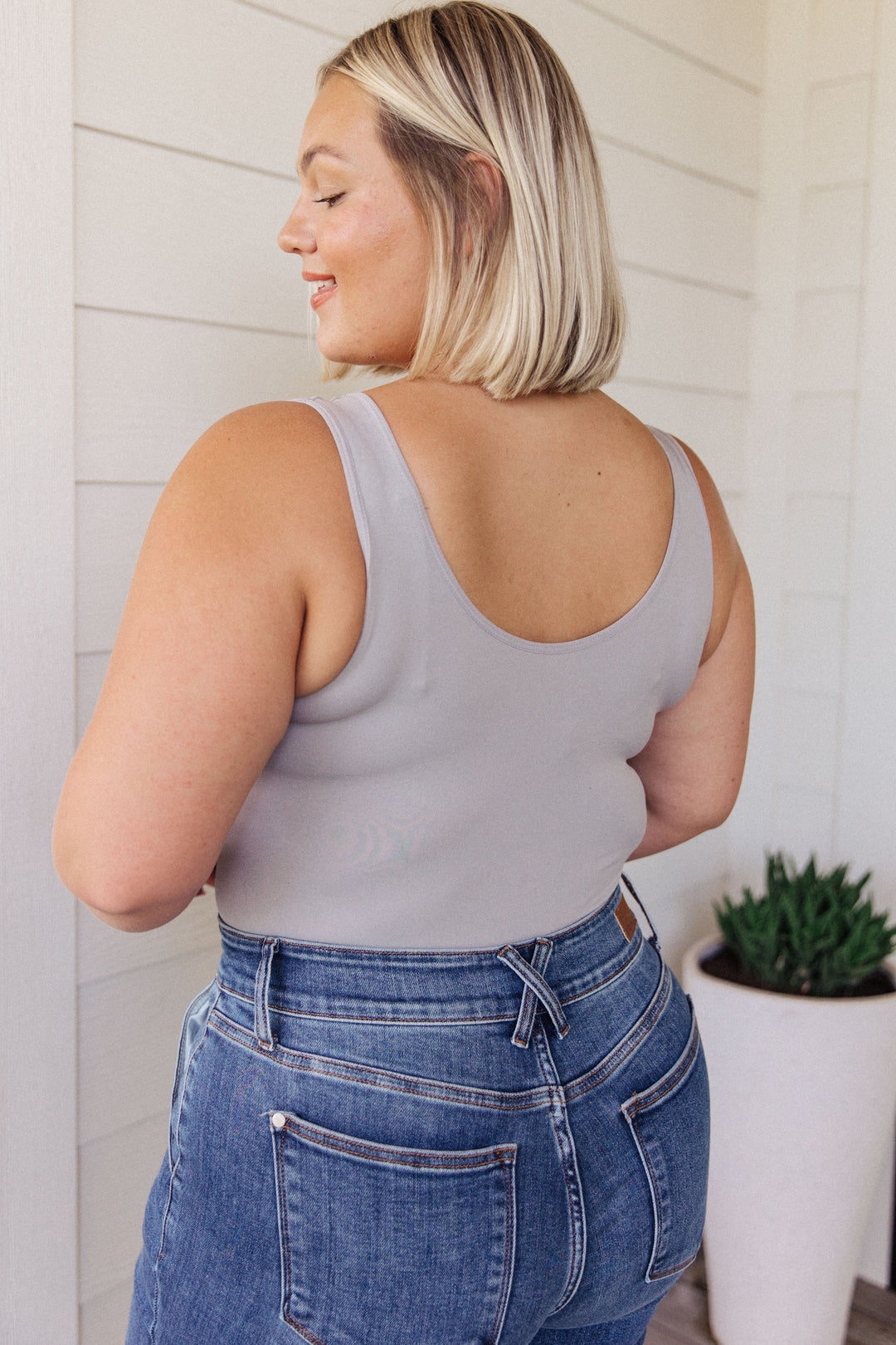 Back view of a plus size model in a gray scoop-neck bodysuit and jeans, standing in front of a white shiplap wall with a potted succulent