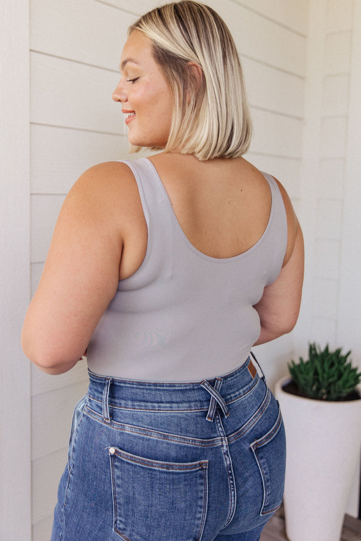 Back view of a plus size model in a gray scoop-neck bodysuit and jeans, standing in front of a white shiplap wall with a potted succulent