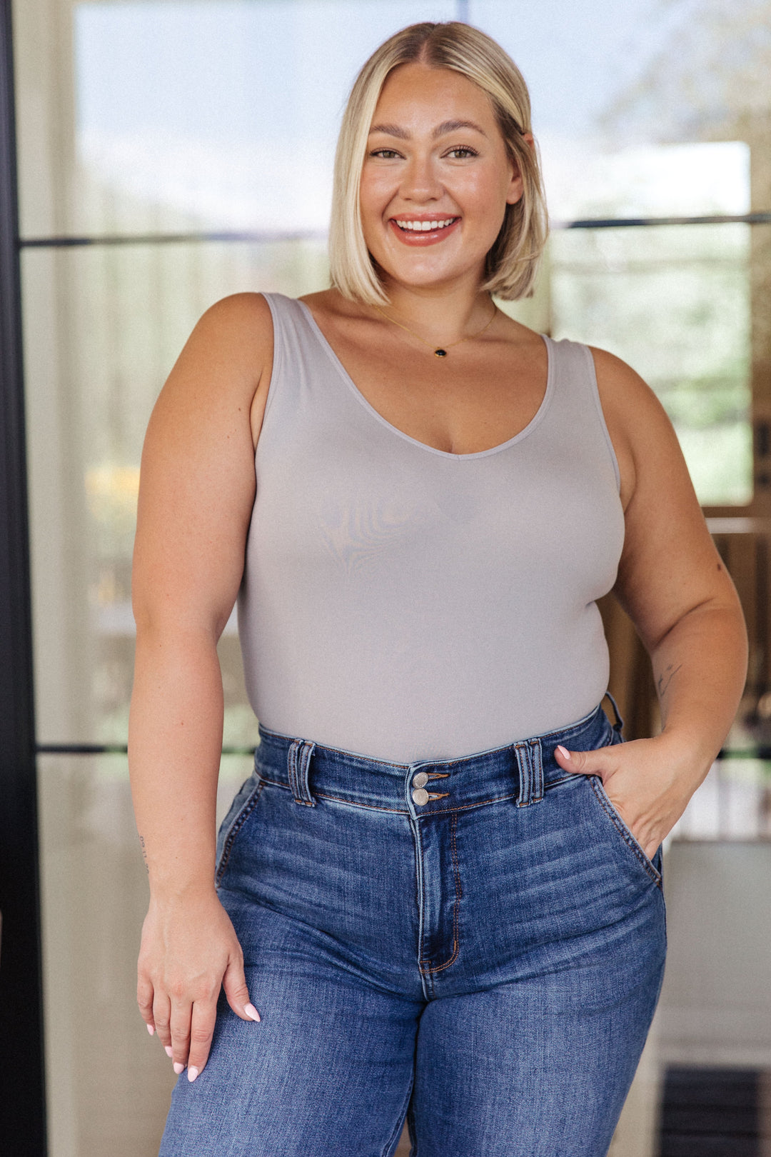 Plus size model smiling in a gray scoop-neck bodysuit and jeans, hand in pocket, standing in front of a glass door