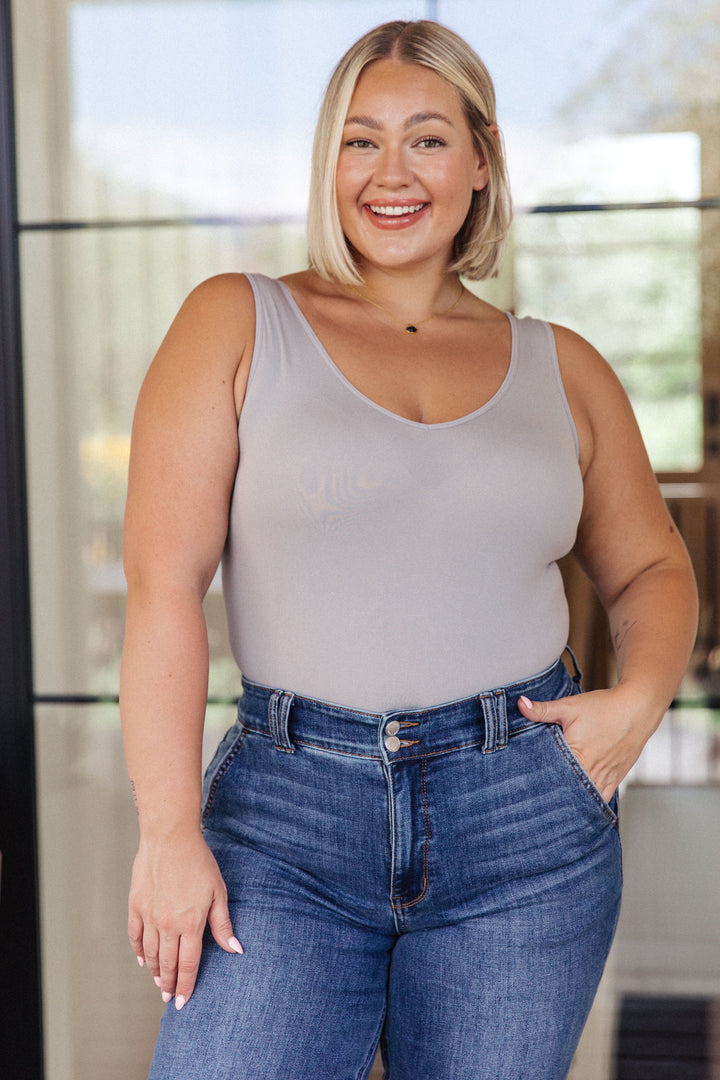 Plus size model smiling in a gray scoop-neck bodysuit and jeans, hand in pocket, standing in front of a glass door