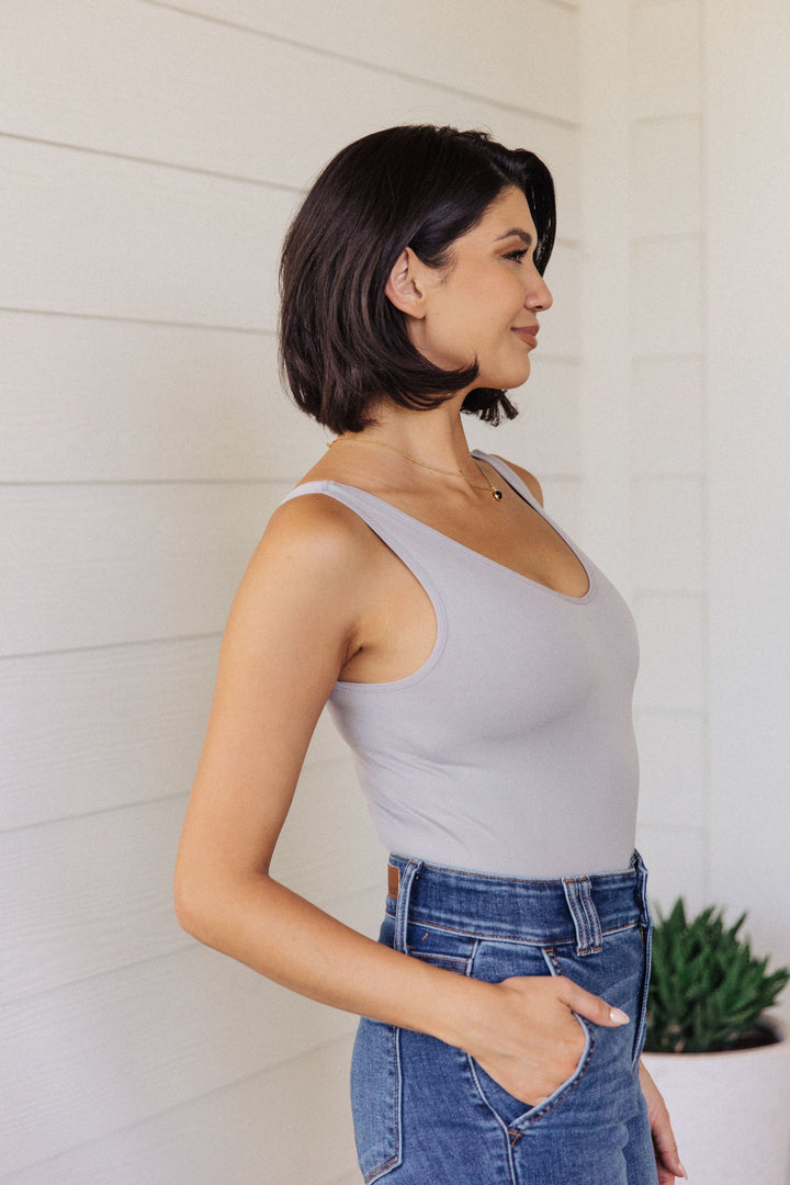 Side view of a woman in a gray scoop-neck bodysuit and jeans, hand in pocket, standing in front of a white shiplap wall with a potted succulent