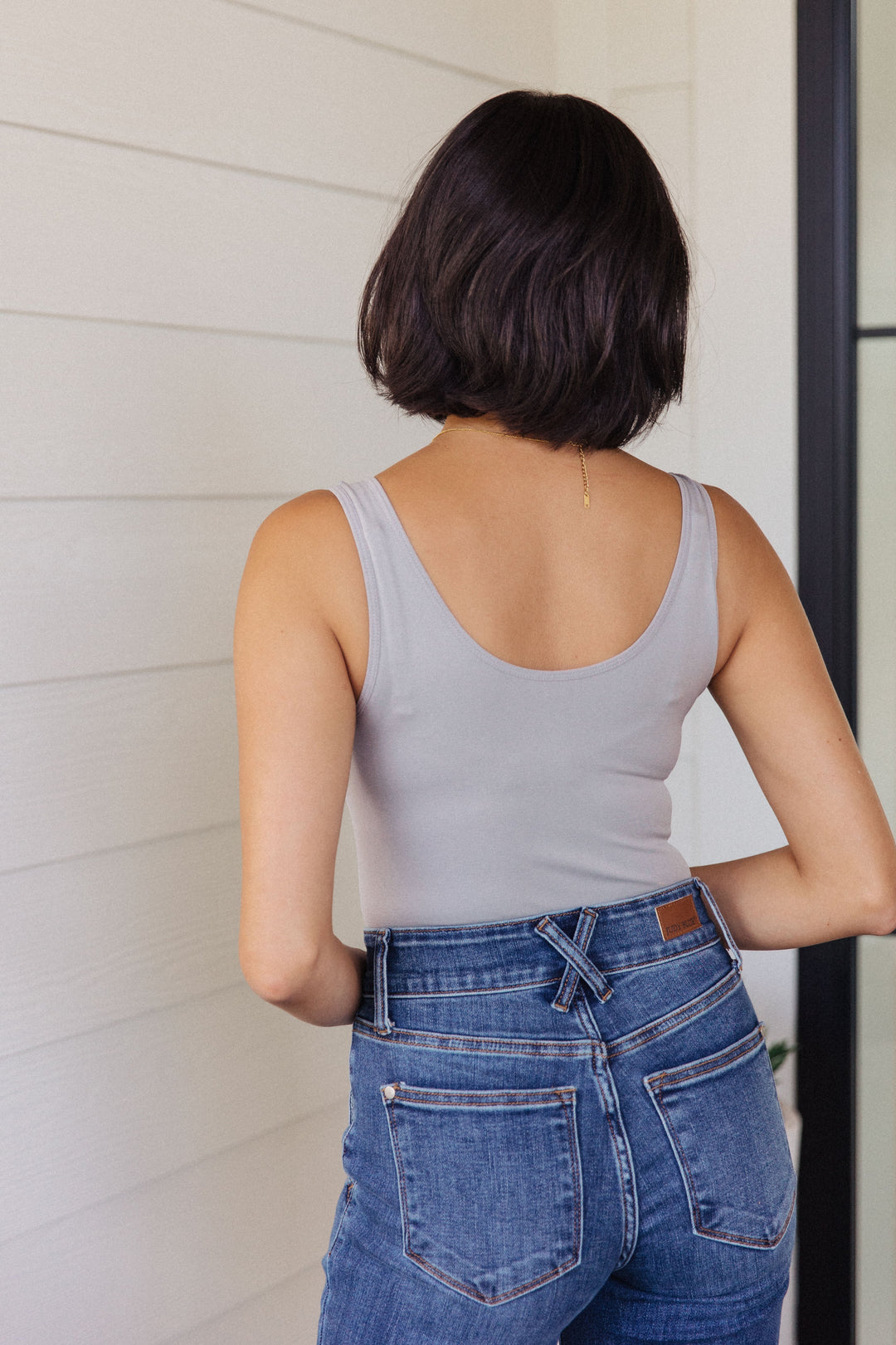 Back view of a woman in a gray scoop-neck bodysuit and jeans, standing in front of a white shiplap wall