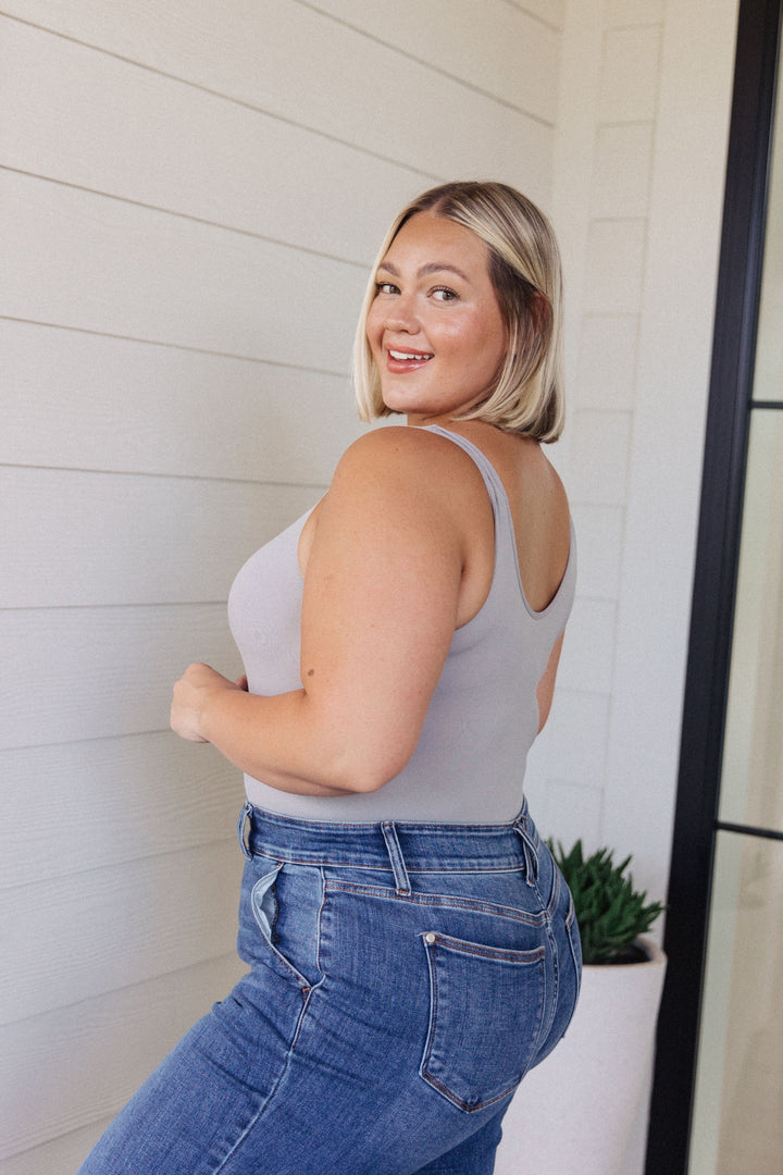 Plus size model smiling over her shoulder in a gray scoop-neck bodysuit and jeans, standing in front of a white shiplap wall with a potted succulent