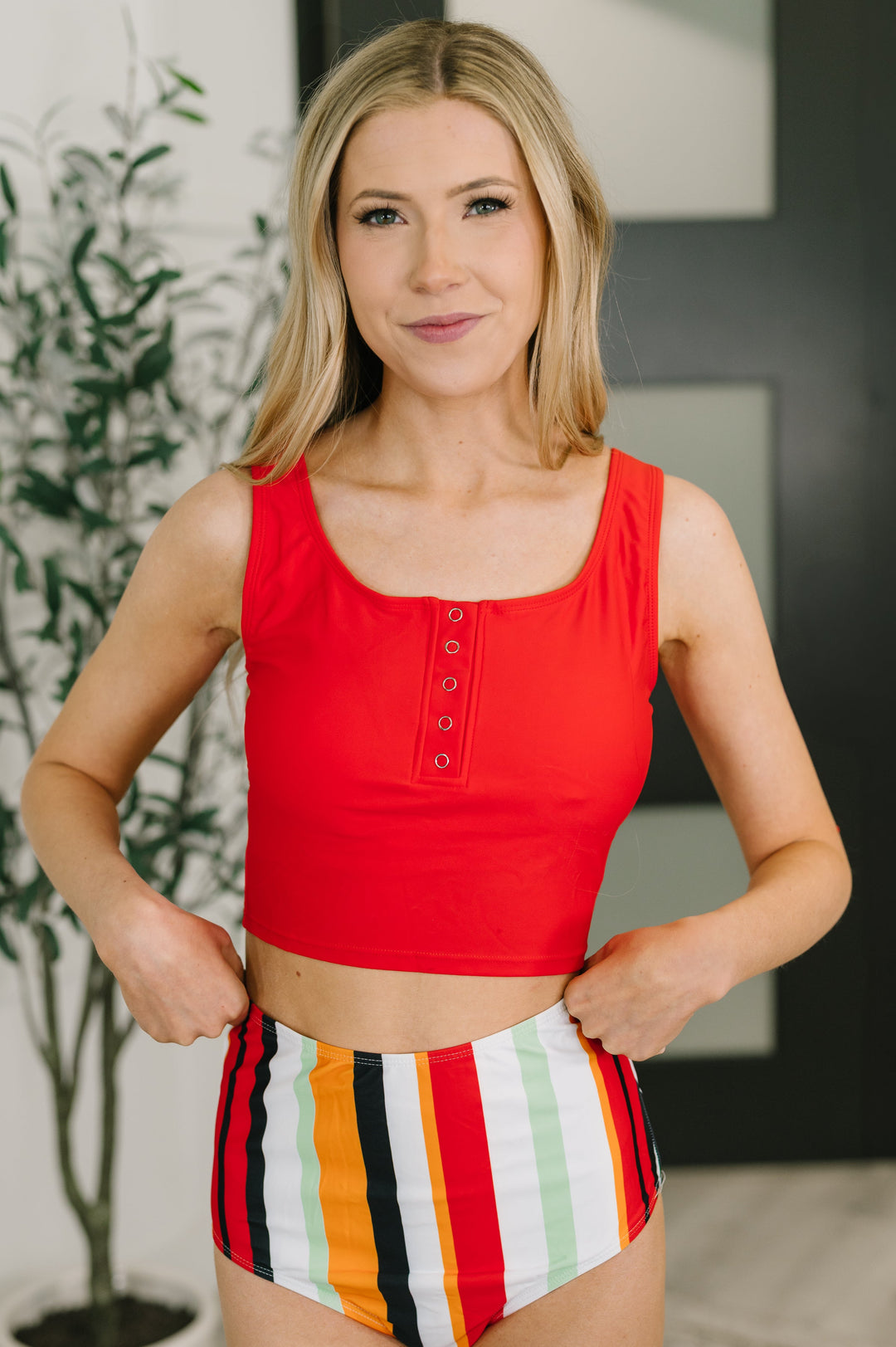 Front view of a woman smiling in a red square-neck button-front crop tank top and high-waist multicolor stripe bikini bottoms, with hands in pockets.