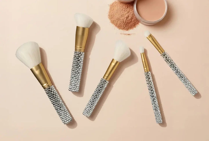 Rhinestone 5-Piece Makeup Brush Set by Genie, flat lay showing all five brushes with crystal-encrusted handles, gold ferrules, and white bristles alongside an open powder compact on a blush background