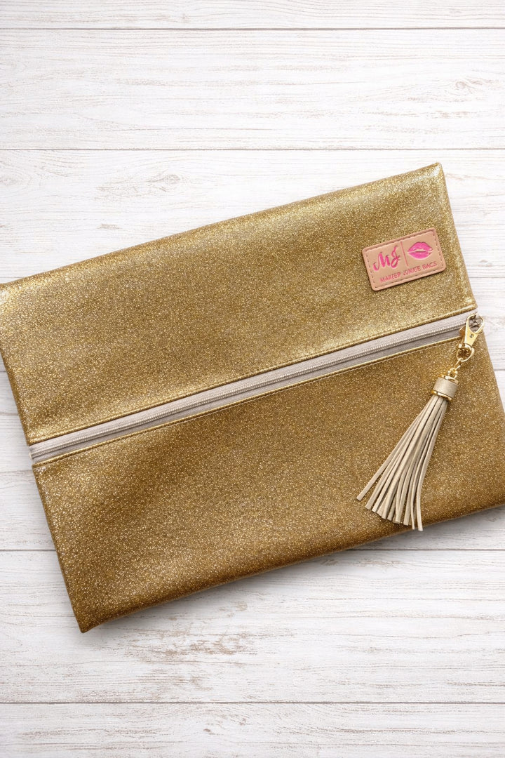 Makeup Junkie Bags gold glitter foldover zip makeup bag with gold tassel zipper pull and branded patch, front view on white wood background