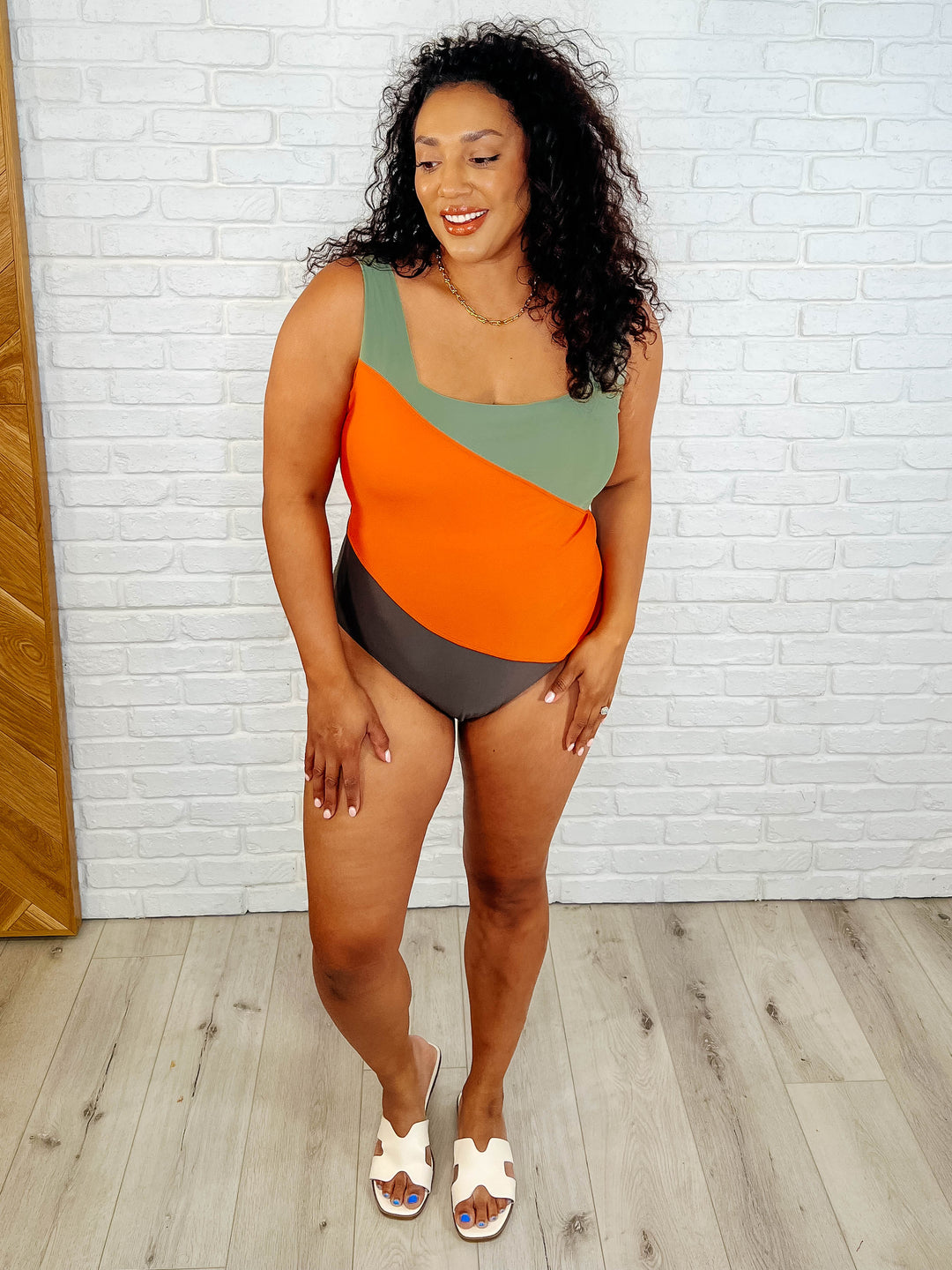Full-length front view of a plus size model smiling in the sage, orange, and dark brown color block one-piece swimsuit, paired with white slide sandals.