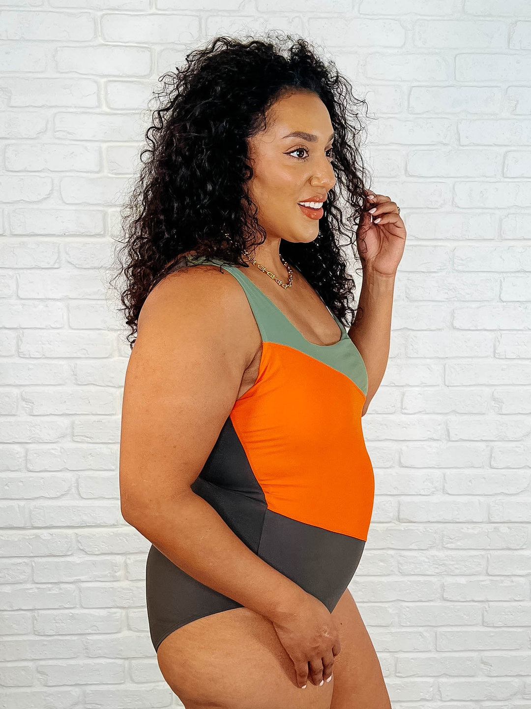 Three-quarter side view of a plus size model smiling in the sage, orange, and dark olive color block one-piece swimsuit.
