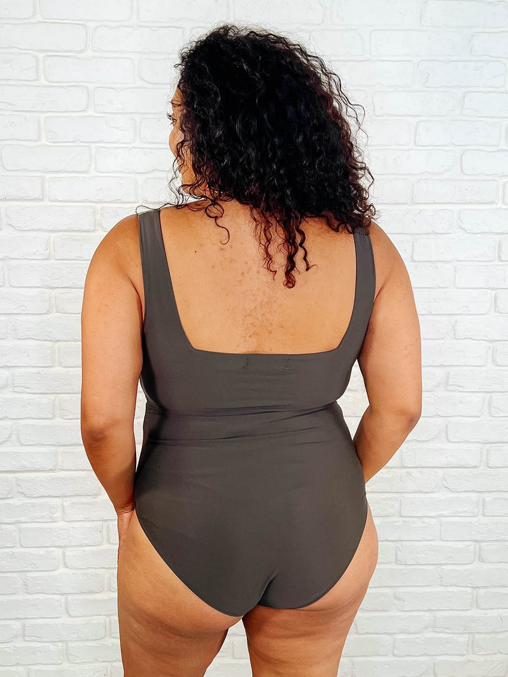 Back view of a plus size model in the dark olive color block one-piece swimsuit with wide square straps.