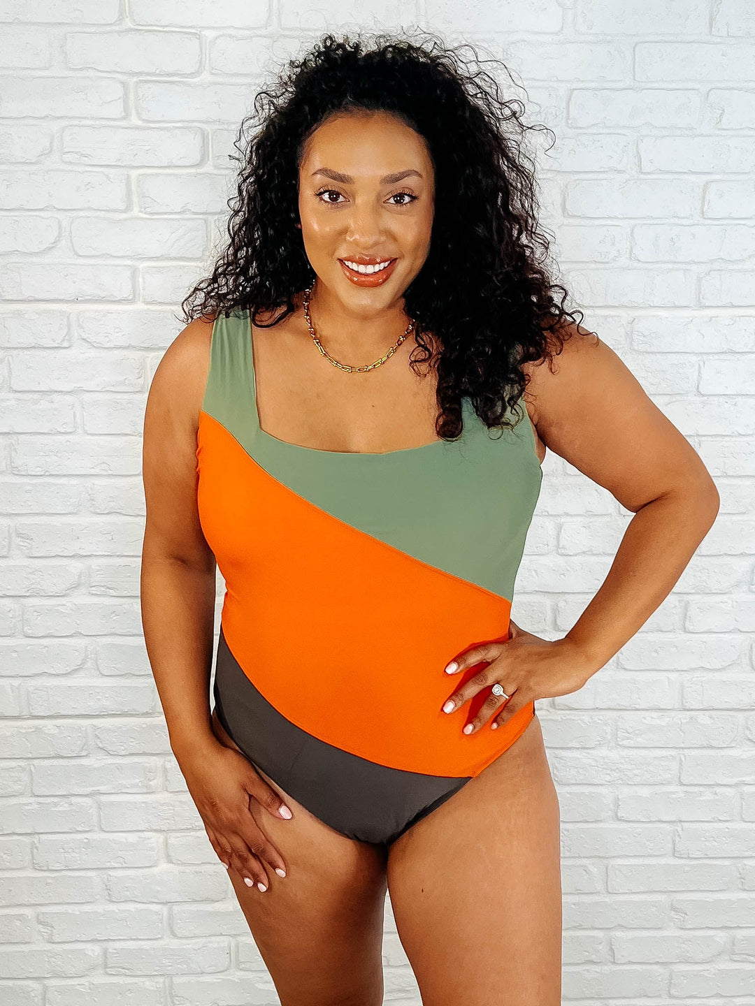 Close-up front view of a plus size model smiling with hand on hip in the sage, orange, and dark brown color block one-piece swimsuit with a gold necklace.