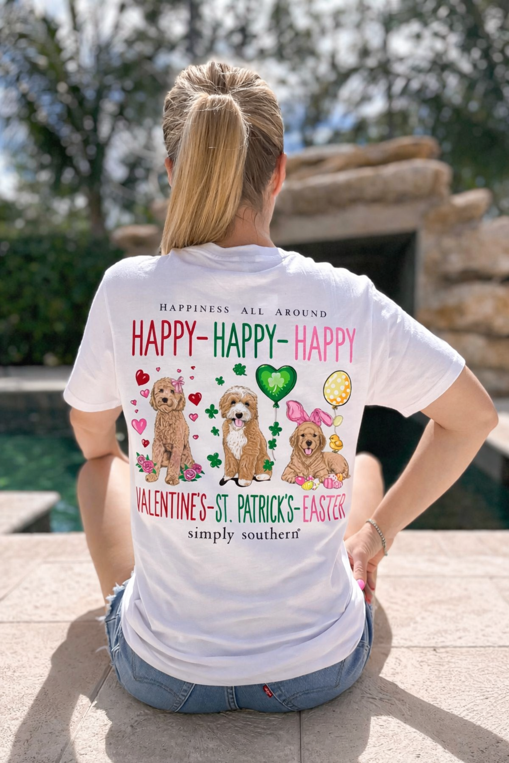 Person wearing a white t-shirt with festive designs and text, sitting by a pool.