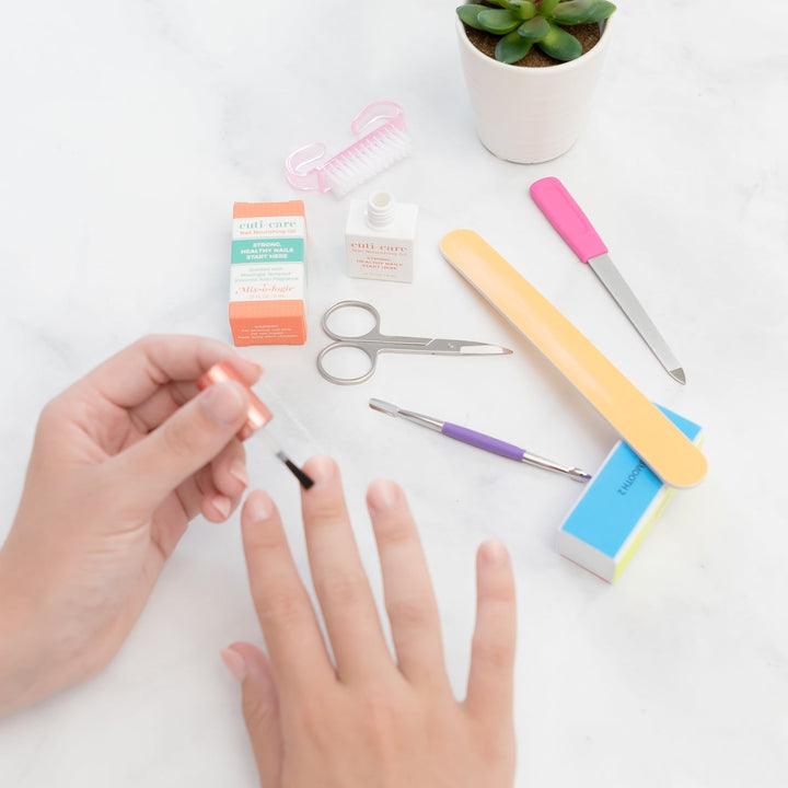 Mixologie Cuti-Care Nail Nourishing Oil being applied with brush applicator on hand, surrounded by nail scissors, file, buffer block, cuticle pusher, and orange box on marble surface with succulent