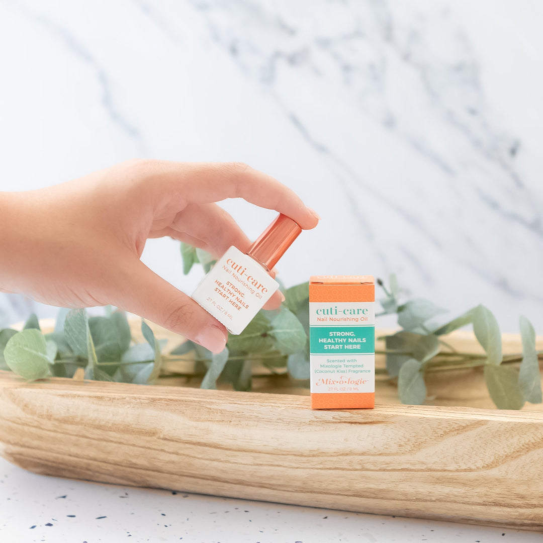 Mixologie Cuti-Care Nail Nourishing Oil, 0.27 fl oz, held in hand over wood tray with eucalyptus and orange box on marble background