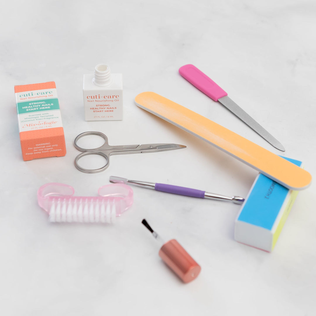 Mixologie Cuti-Care Nail Nourishing Oil flat lay with orange box, white bottle, nail scissors, emery board, buffer block, cuticle pusher, and pink toe separator on white marble background