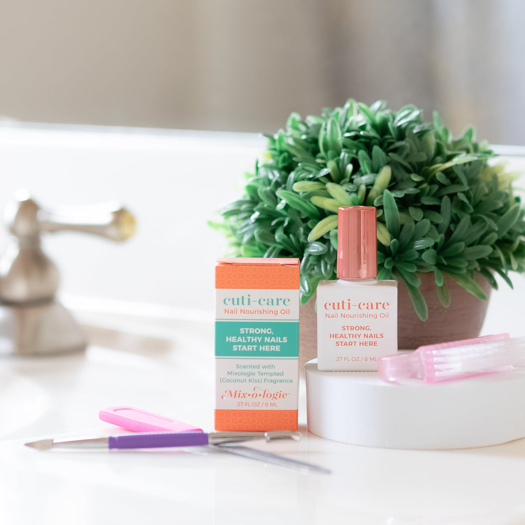 Mixologie Cuti-Care Nail Nourishing Oil, white bottle with rose gold cap and orange box styled on bathroom counter with greenery, pink scissors, and cuticle pusher
