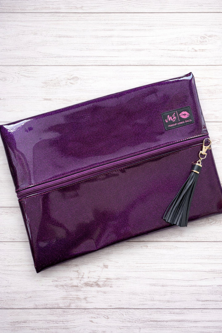 Makeup Junkie Bags deep purple glitter foldover zip makeup bag with navy tassel zipper pull and branded patch, front view on white wood background