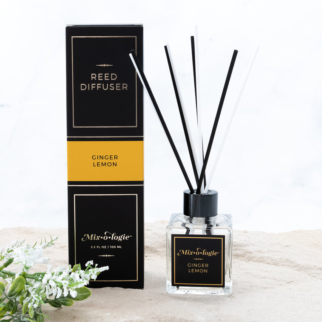 Mixologie Reed Diffuser in Ginger Lemon, 3.3 fl oz, clear square glass bottle with black cap and black label with gold accent band, with matching black and gold box, styled with white florals