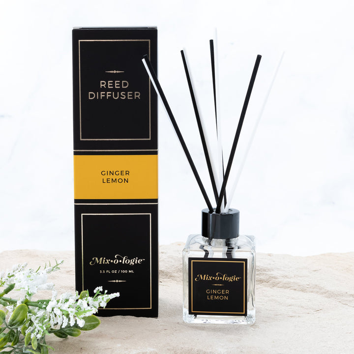 Mixologie Reed Diffuser in Ginger Lemon, 3.3 fl oz, clear square glass bottle with black cap and black label with gold accent band, with matching black and gold box, styled with white florals