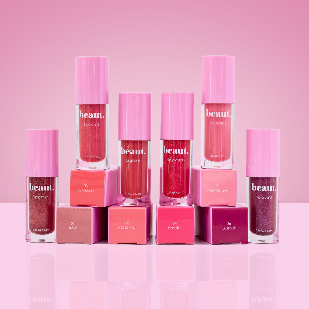 Beaut. Lip Gloss'd six shades in clear tubes with pink caps displayed with color-matched square boxes in shades 01 Girly Guava, 02 Blossom'd, 03 Sun Kiss'd, 04 Bashful, 05 Spice, and 06 Blush'd on pink background