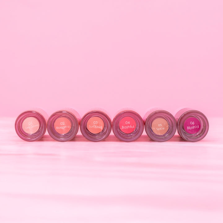 Beaut. Lip Gloss'd six shades in clear tubes with pink caps displayed bottom-up showing shade names: 01 Girly Guava, 02 Blossom'd, 03 Sun Kiss'd, 04 Bashful, 05 Spice, and 06 Blush'd on pink background