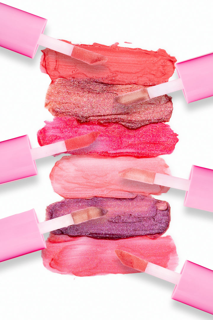 Beaut. Lip Gloss'd color swatches in six shades with pink-capped applicators showing coral red, rose gold glitter, hot pink, blush, purple glitter, and pink on white background