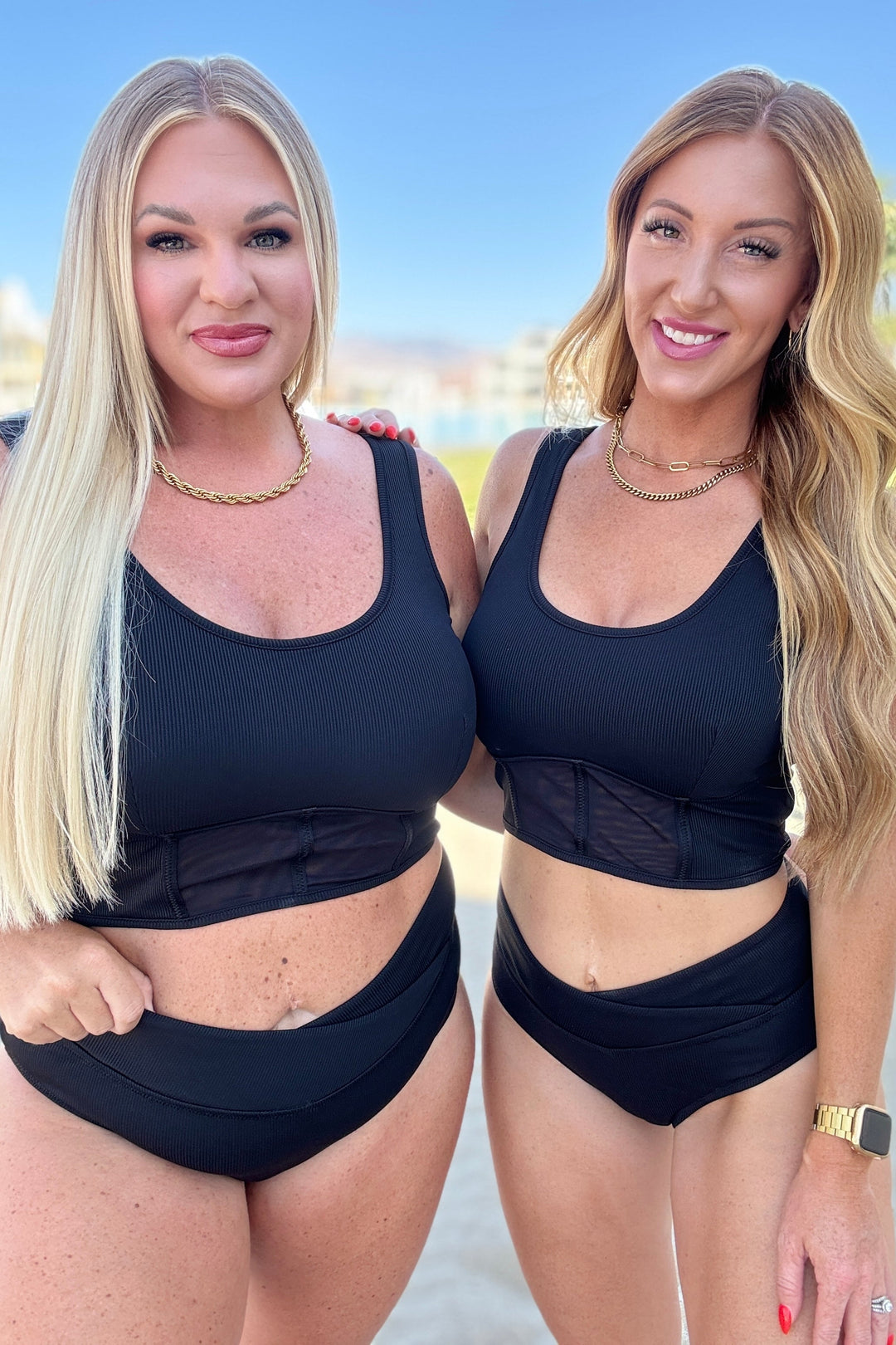 Two women wearing matching black ribbed crop bikini tops with mesh waist panels and high-waist V-front bikini bottoms, styled with gold chain necklaces outdoors.