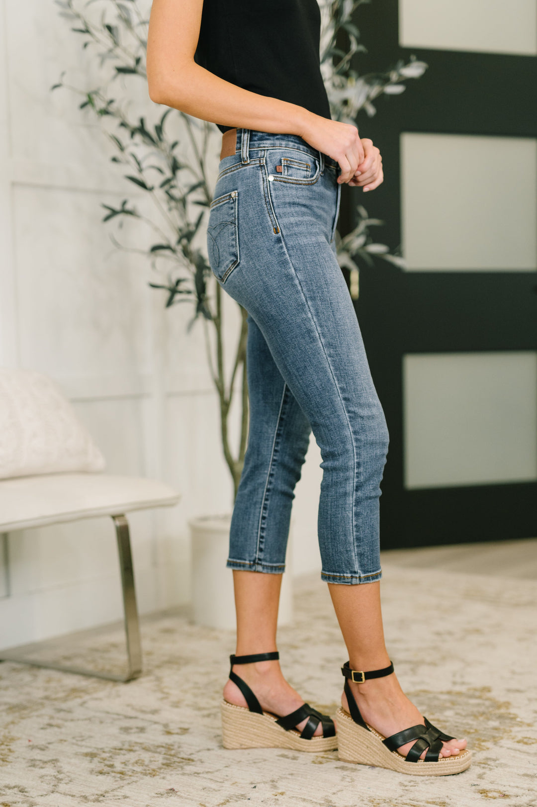 Side view of model wearing Judy Blue medium wash high-rise tummy control rhinestone letter-print cropped straight jeans, styled with a black tank top and black espadrille wedge sandals.