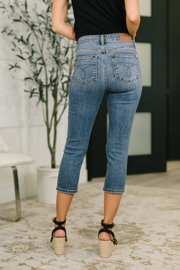 Back view of model wearing Judy Blue medium wash high-rise tummy control rhinestone letter-print cropped straight jeans, styled with a black tank top and black espadrille wedge sandals.