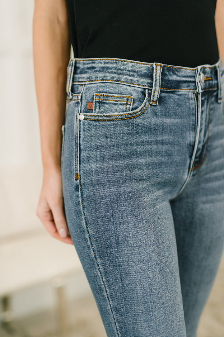 Close-up of Judy Blue medium wash high-rise cropped jeans showing rhinestone letter-print fabric texture, coin pocket, and leather brand patch detail.
