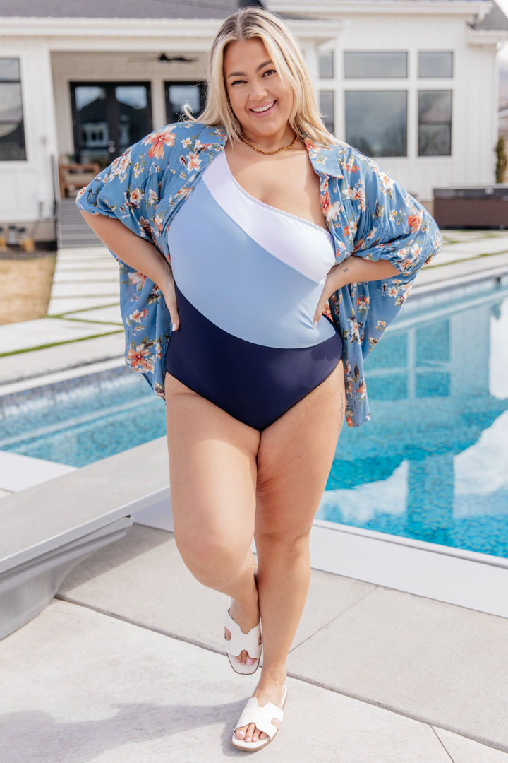 Plus size model wearing a white, light blue, and navy colorblock one-shoulder one-piece swimsuit layered under an open blue floral kimono cover-up, styled with white slide sandals poolside.