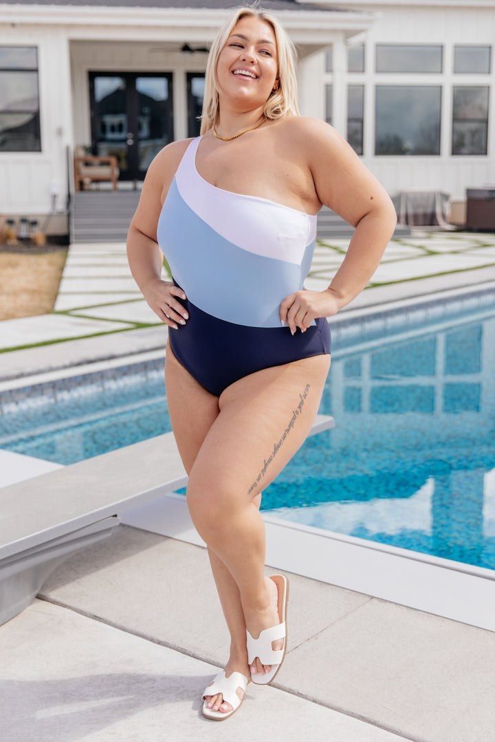 Plus size model wearing a white, light blue, and navy colorblock one-shoulder one-piece swimsuit, styled with white slide sandals and a gold chain necklace poolside.