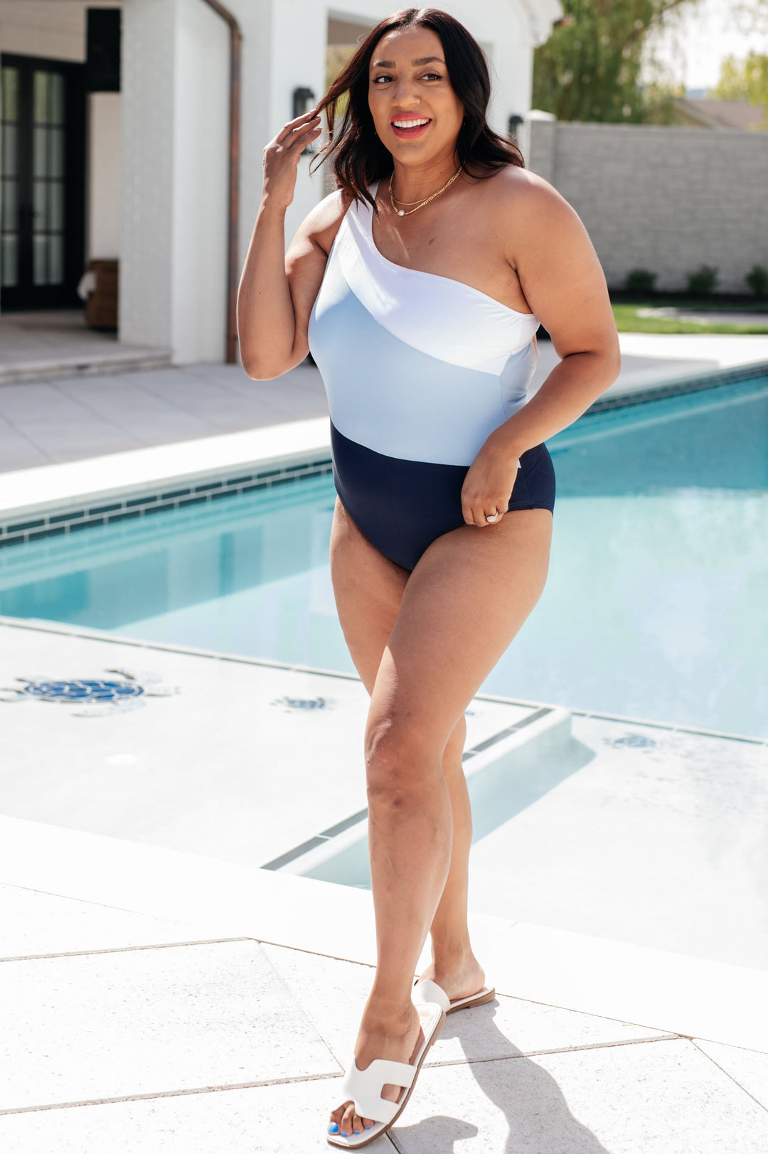 Plus size model wearing a white, light blue, and navy colorblock one-shoulder one-piece swimsuit, styled with white slide sandals and a gold chain necklace poolside.