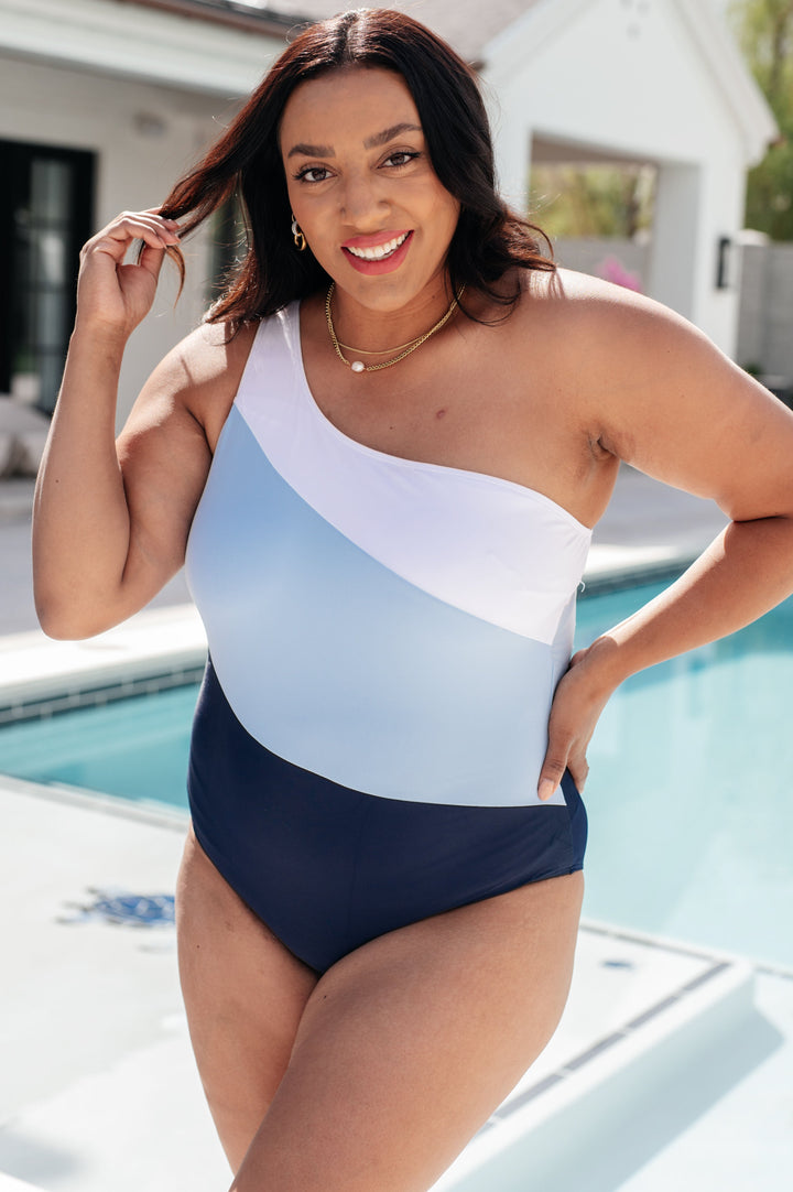 Plus size model wearing a white, light blue, and navy colorblock one-shoulder one-piece swimsuit, styled with gold hoop earrings and a layered gold necklace poolside.