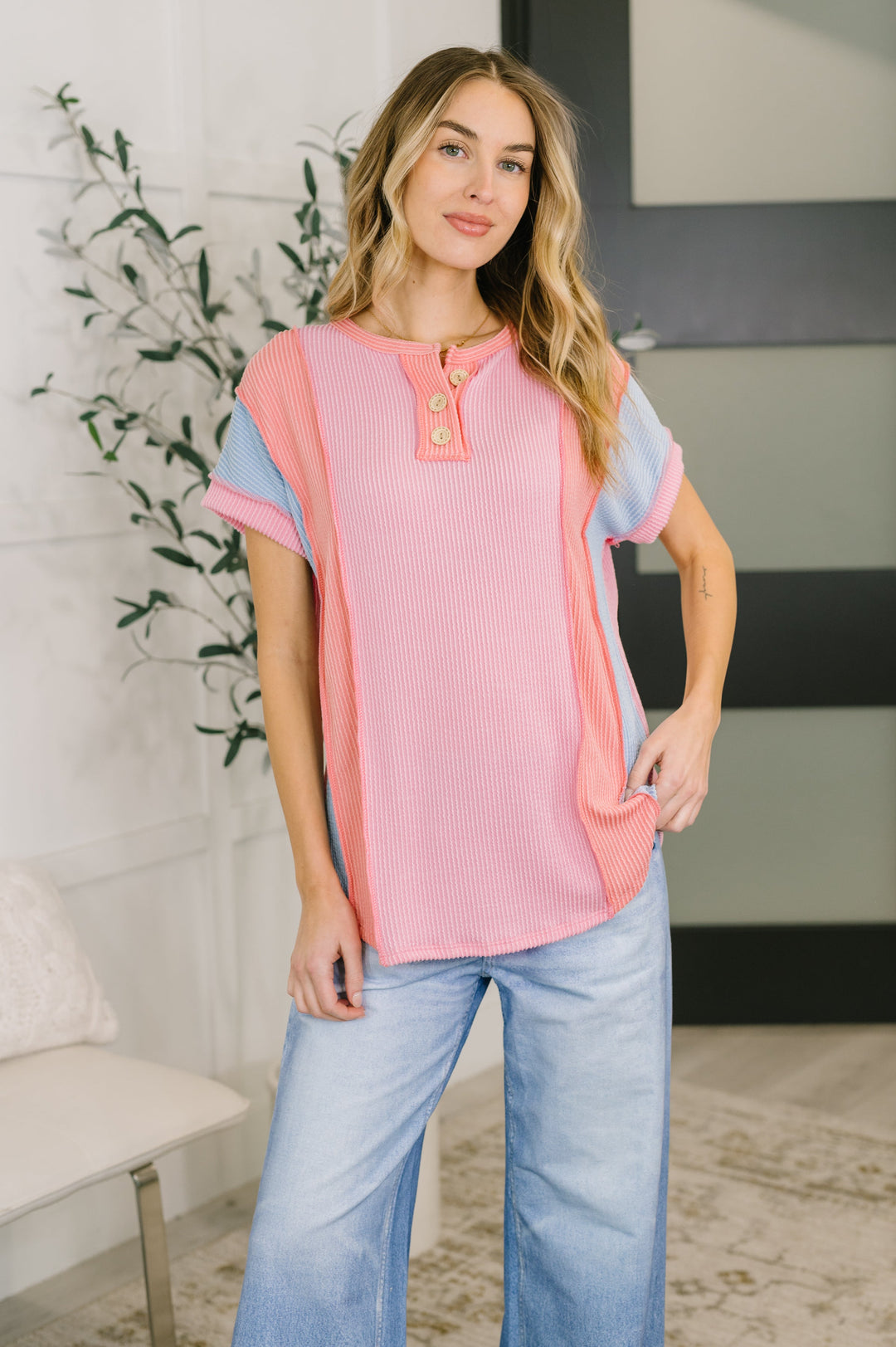 Woman wearing a pink and blue color block short-sleeve henley top with wide-leg jeans, posed in front of a white bench and leafy plant