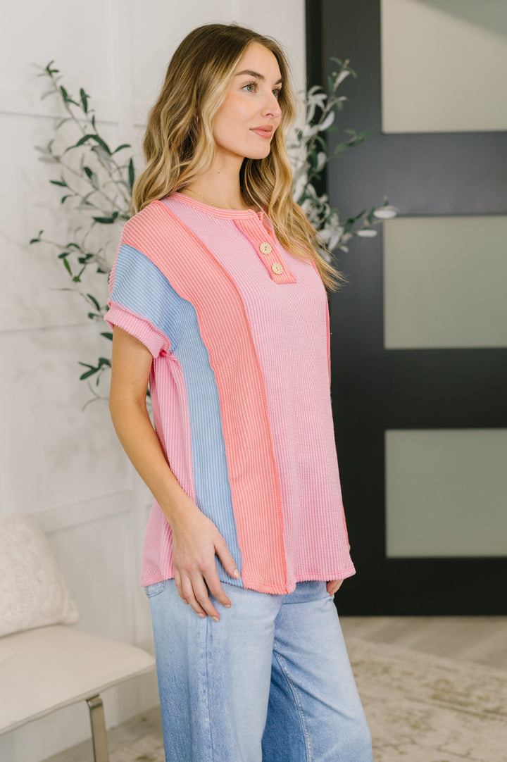 Side view of a woman wearing a pink and blue color block short-sleeve henley top with wide-leg jeans, posed in front of a white bench and leafy plant