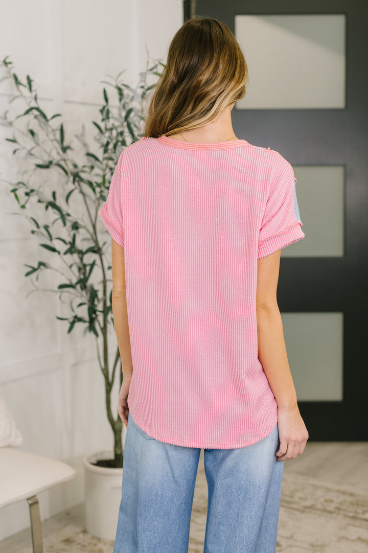 Back view of a woman wearing a pink ribbed short-sleeve top with wide-leg jeans, posed in front of a white bench and leafy plant