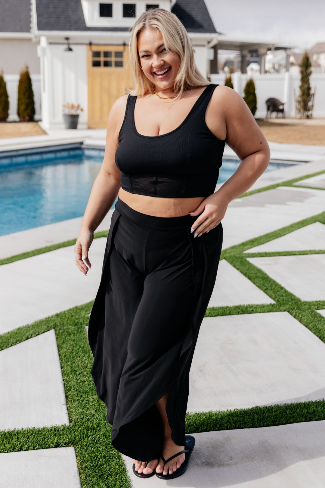 Plus size model wearing a black scoop neck crop bikini top and black wide-leg maxi skirt cover-up with front slit, styled with black flip flops poolside.