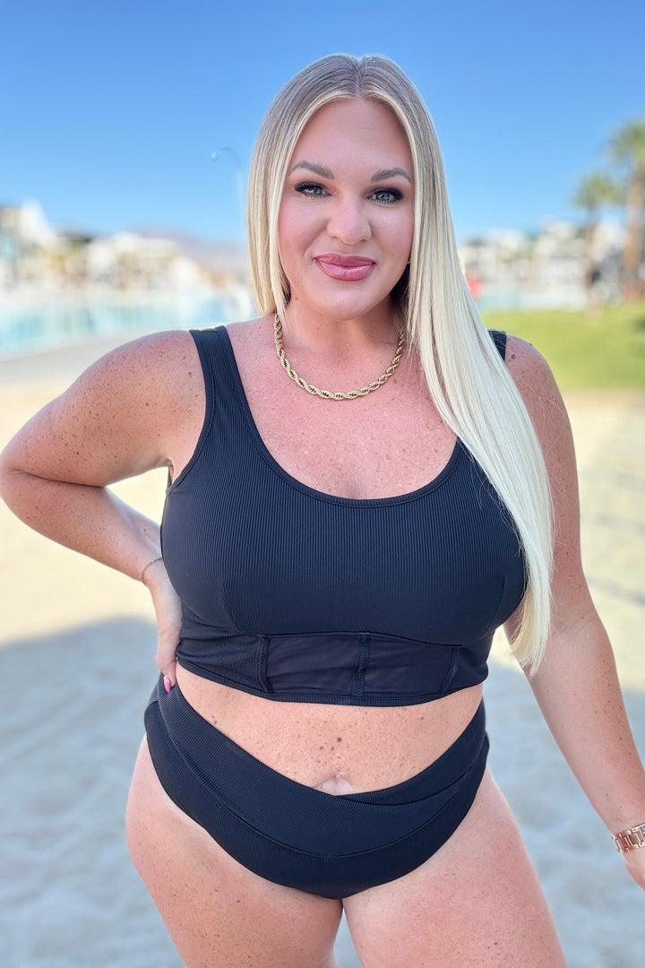 Plus size model wearing a black ribbed crop bikini top with mesh waist panel and matching high-waist bikini bottoms with V-front detail, styled with a gold chain necklace outdoors.
