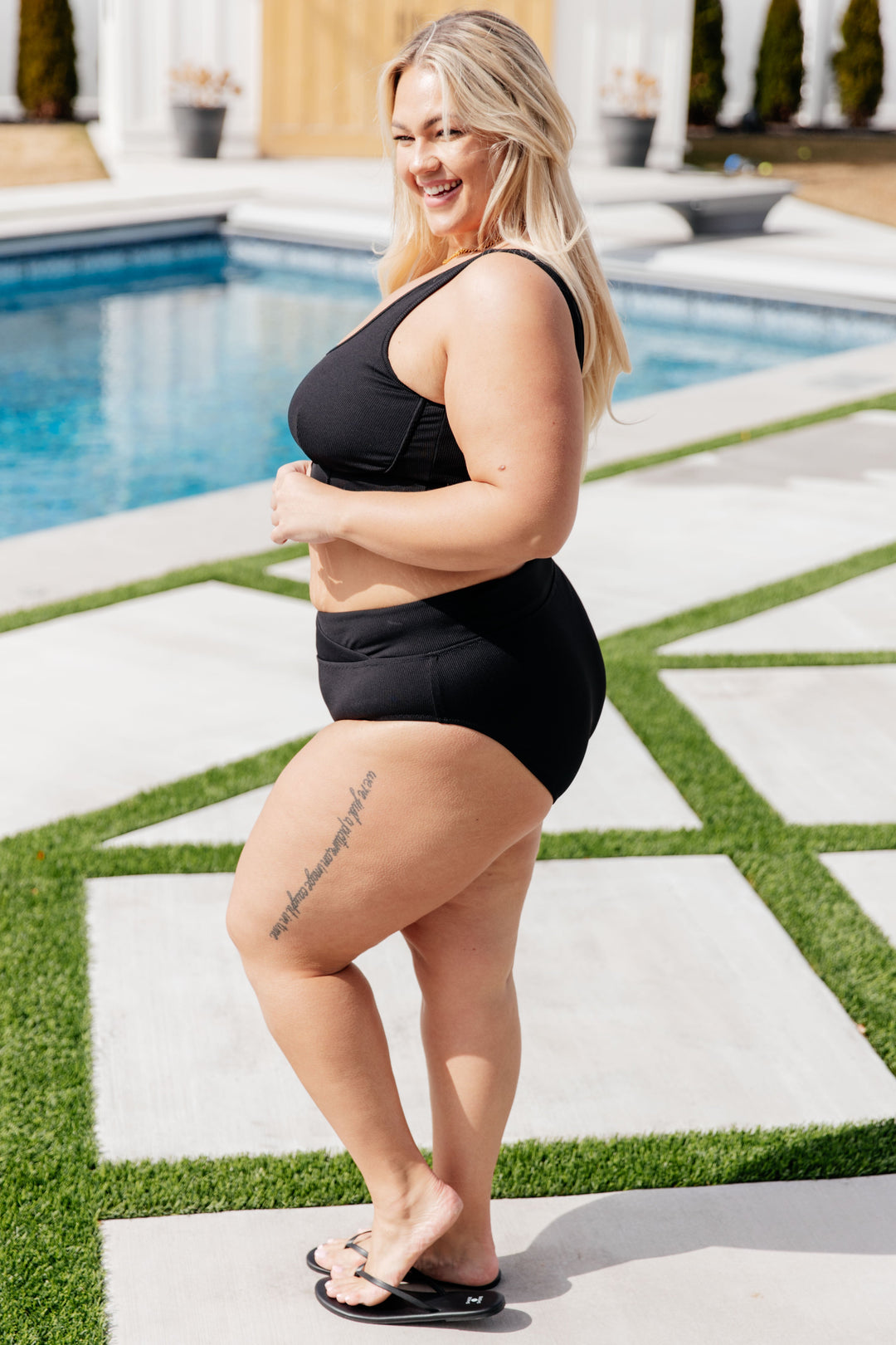 Side view of plus size model wearing a black ribbed crop bikini top and high-waist bikini bottoms, styled with black flip flops poolside.